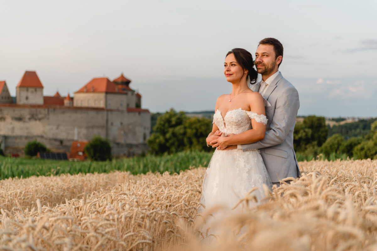 Ein Paar im Hochzeitskleid steht bei Sonnenuntergang in einem Weizenfeld mit einem historischen Schloss im Hintergrund.