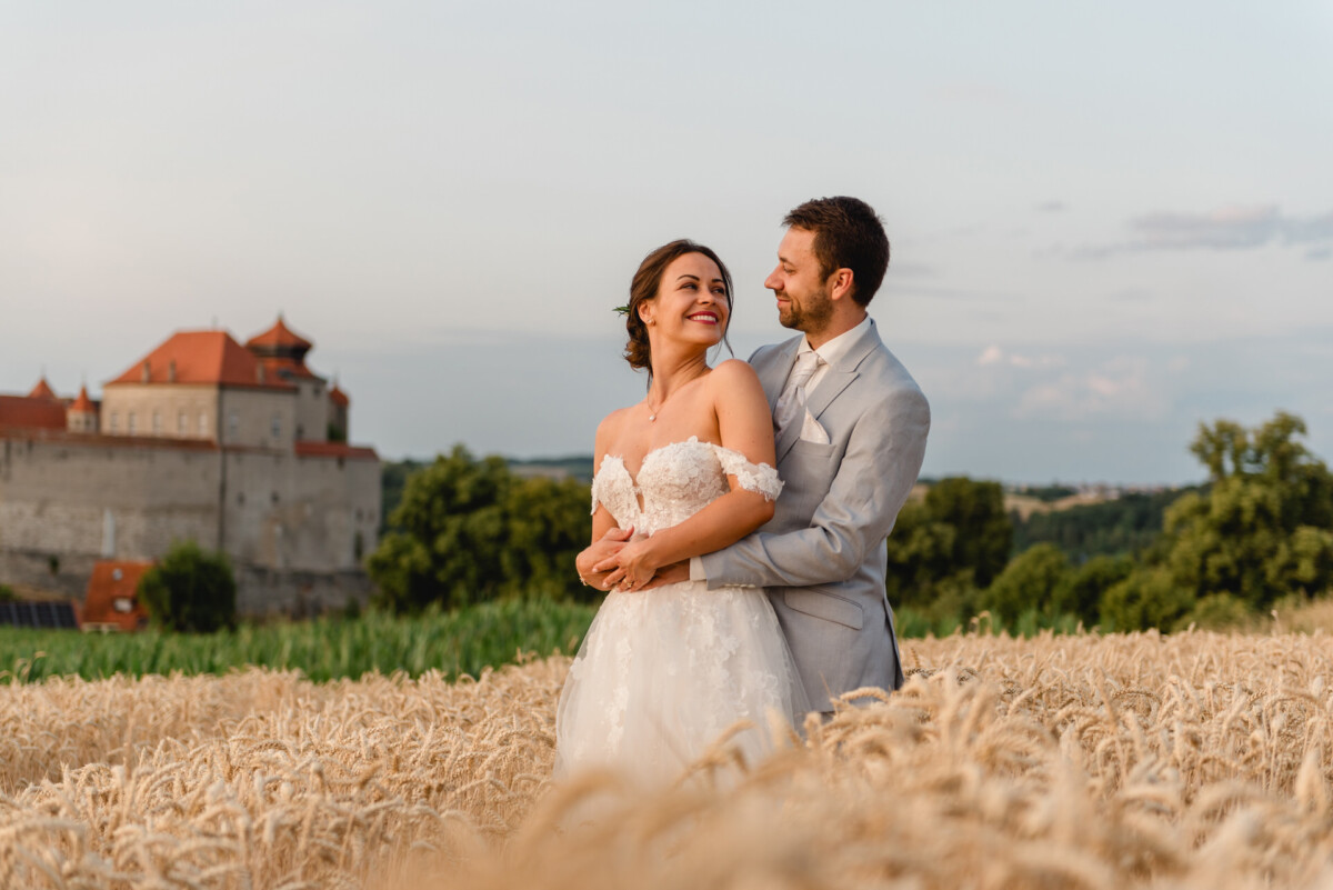 Ein Paar im Hochzeitskleid umarmt sich in einem Weizenfeld, im Hintergrund ist ein Schloss zu sehen.