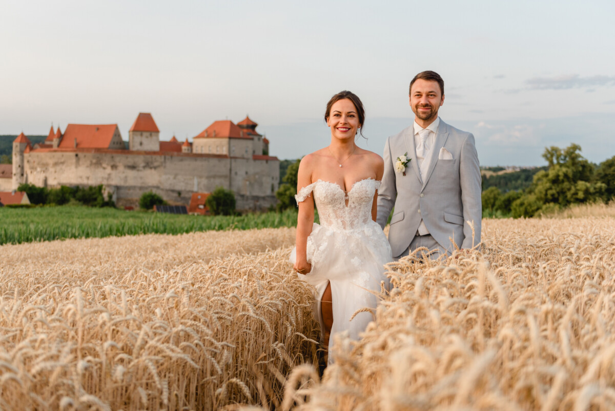 Braut und Bräutigam gehen durch ein Weizenfeld mit einem Schloss im Hintergrund.