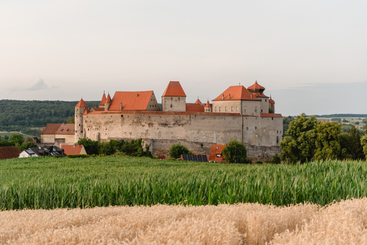 Eine mittelalterliche Burg mit roten Ziegeldächern und Steinmauern steht umgeben von Feldern und Grün unter einem klaren Himmel.