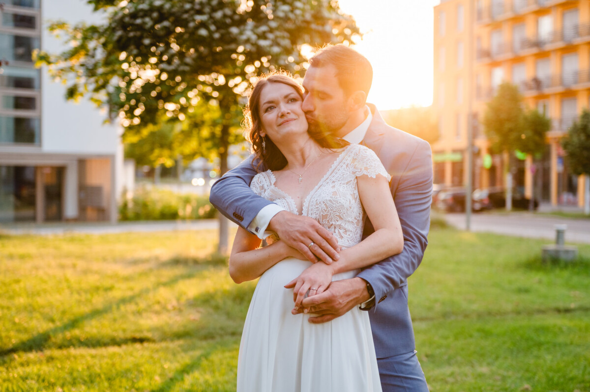 S_S_Hochzeit_Standesamt_Friedberg_Augsburg_nuno_00808 Eine Braut und ein Bräutigam umarmen sich vor einem Gebäude bei Sonnenuntergang.