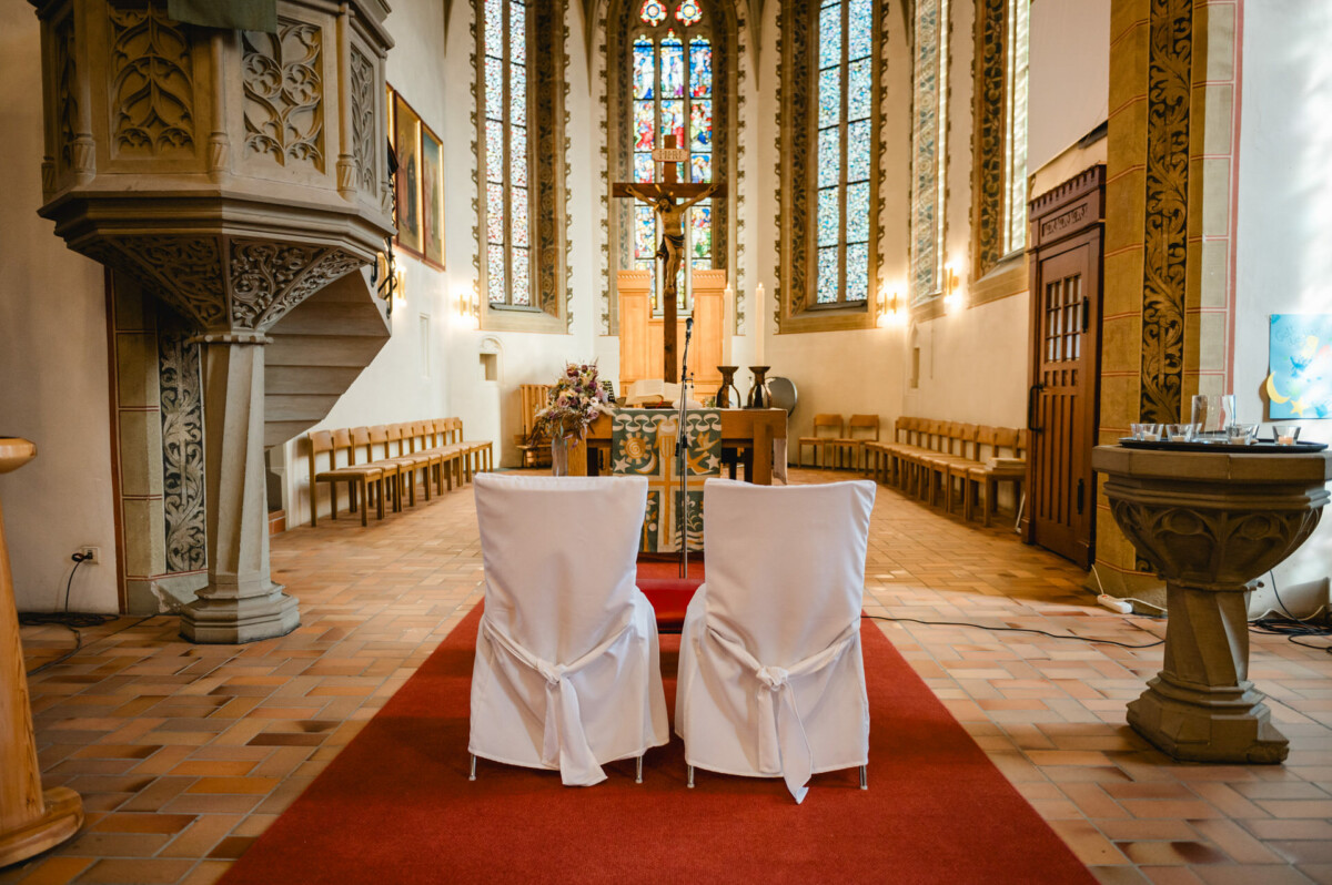 Zwei weiße Stühle stehen vor dem Altar in einer Kirche mit kunstvollen Buntglasfenstern, einer holzgeschnitzten Kanzel und einem Gang mit rotem Teppich.