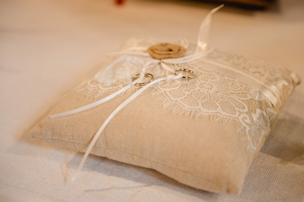 Auf einem beigefarbenen Kissen mit Spitze befinden sich zwei silberne Ringe, die mit weißen Bändern zusammengebunden sind.
