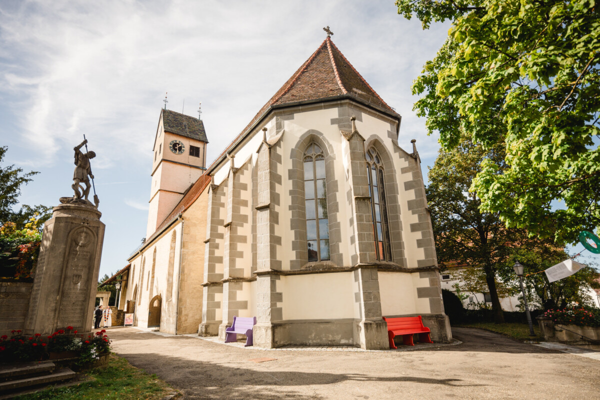 Historische Kirche mit hohem Glockenturm, umgeben von Bäumen und Statuen. Draußen stehen eine rote und eine violette Bank. Schatten deuten auf einen sonnigen Tag hin.