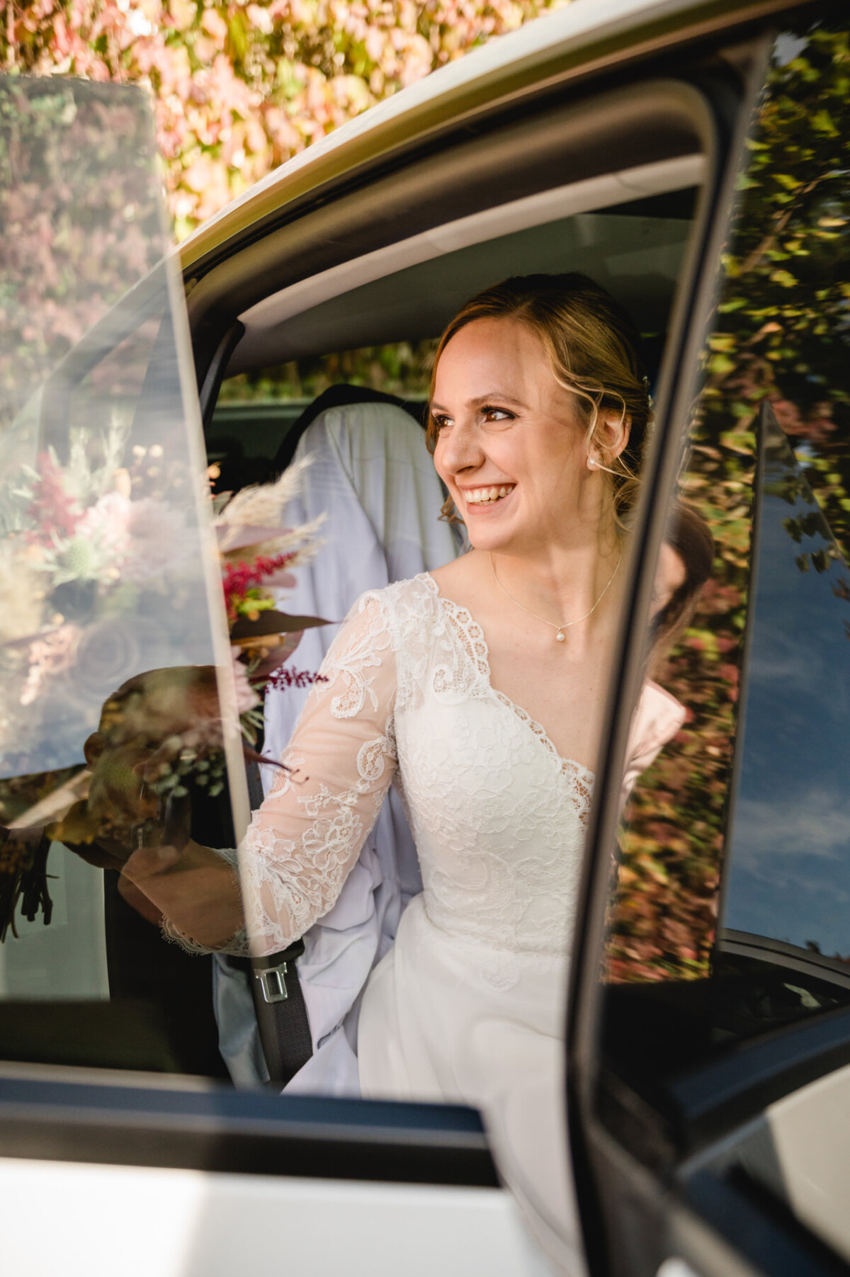 Eine Braut in einem Spitzenbrautkleid sitzt in einem Auto, hält einen Blumenstrauß und lächelt.