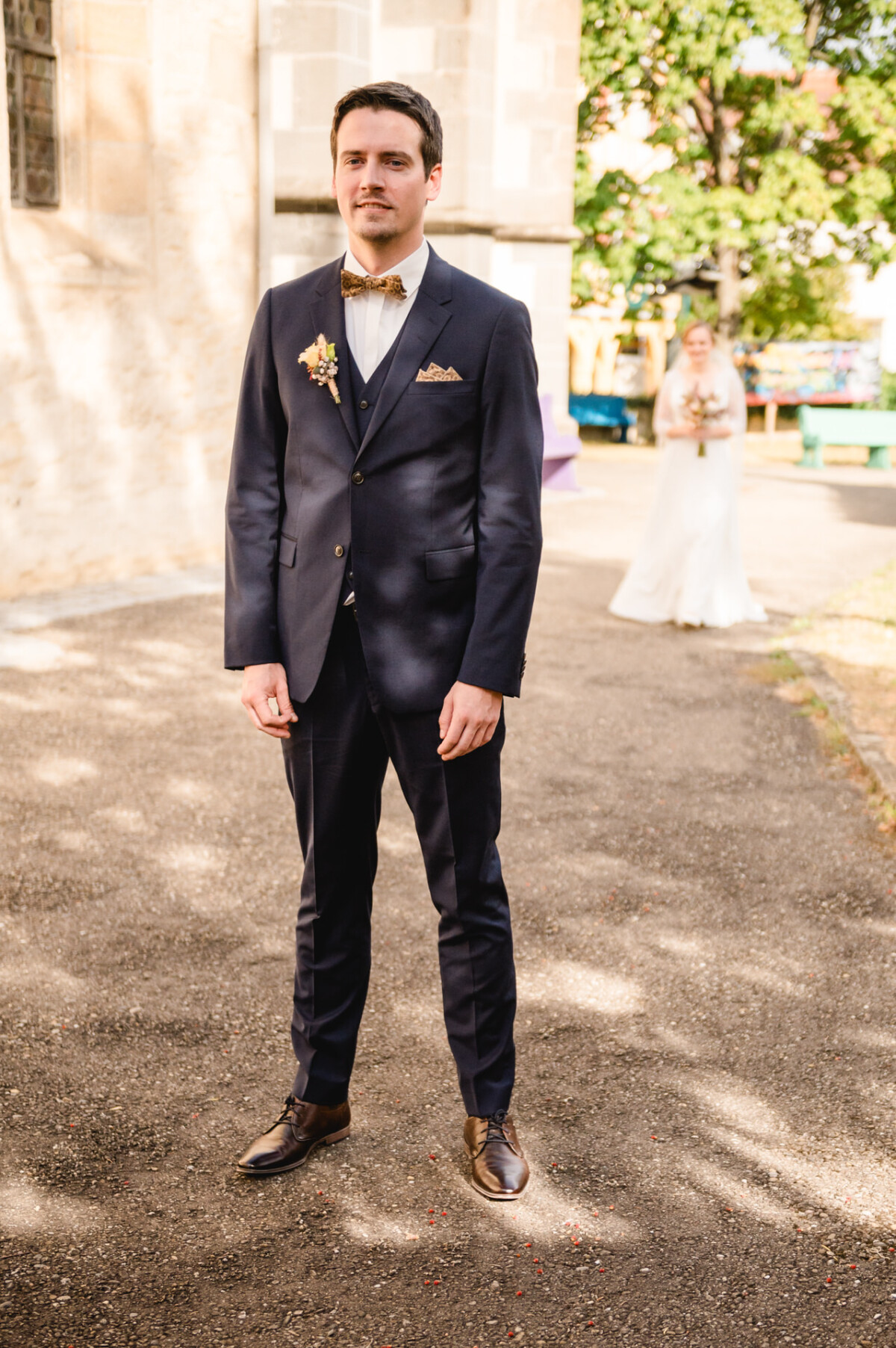 An einem sonnigen Tag steht ein Mann im Anzug im Freien, im Hintergrund hält eine Frau im Brautkleid einen Blumenstrauß.