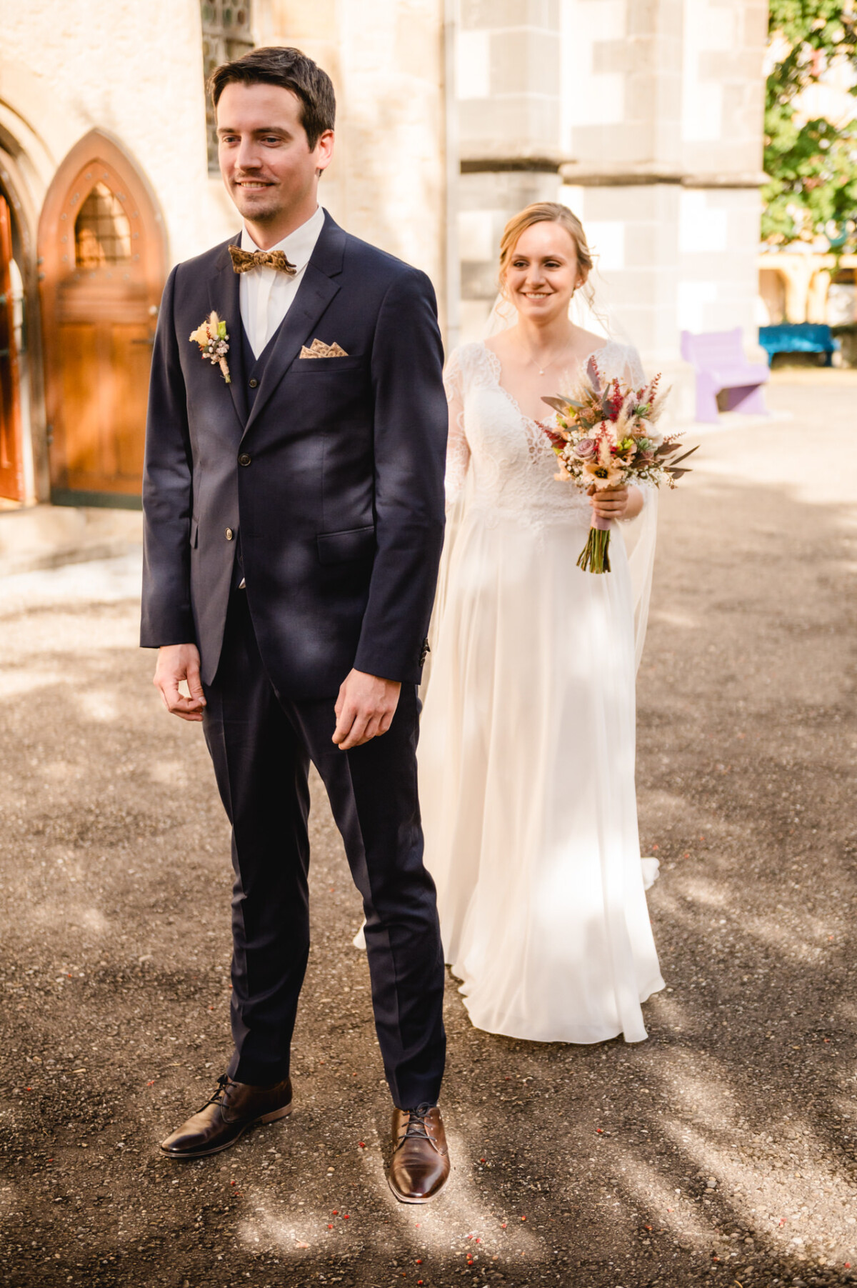 Eine Braut in einem weißen Kleid und ein Bräutigam in einem dunklen Anzug stehen vor einem Gebäude. Die Braut hält einen Blumenstrauß und lächelt, während der Bräutigam nach vorne blickt.