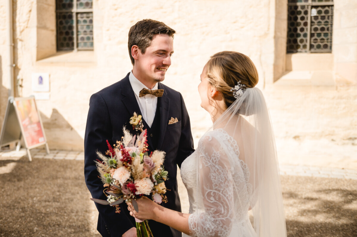 Draußen lächeln sich Braut und Bräutigam an. Die Braut hält einen Blumenstrauß in der Hand. Sie stehen in der Nähe eines Steingebäudes.