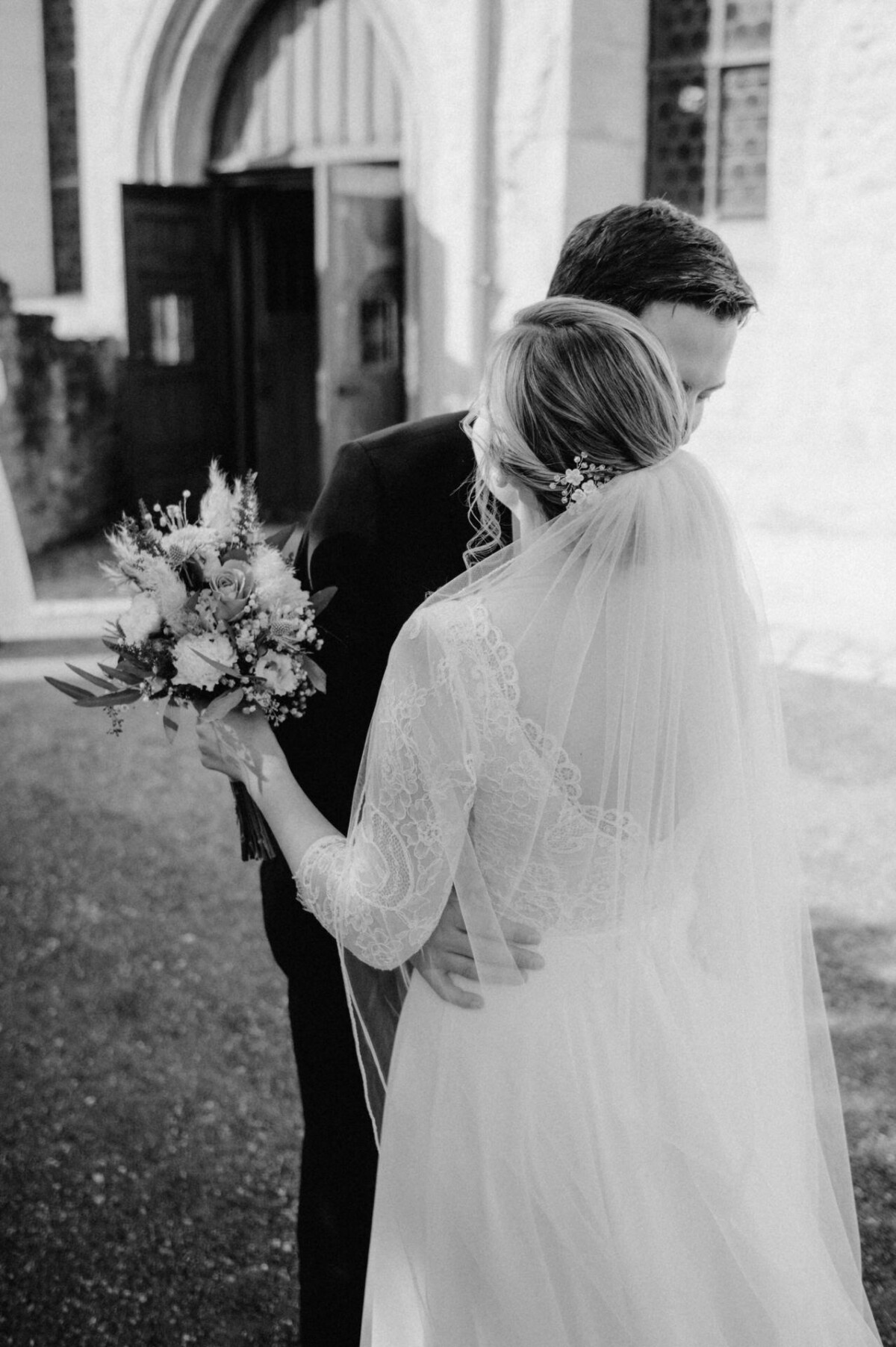 Braut und Bräutigam umarmen sich vor einer Kirche. Die Braut hält einen Blumenstrauß in der Hand, ihr Schleier fällt ihr über den Rücken. Der Bräutigam hält sanft ihre Taille. Im Hintergrund steht die Kirchentür offen.