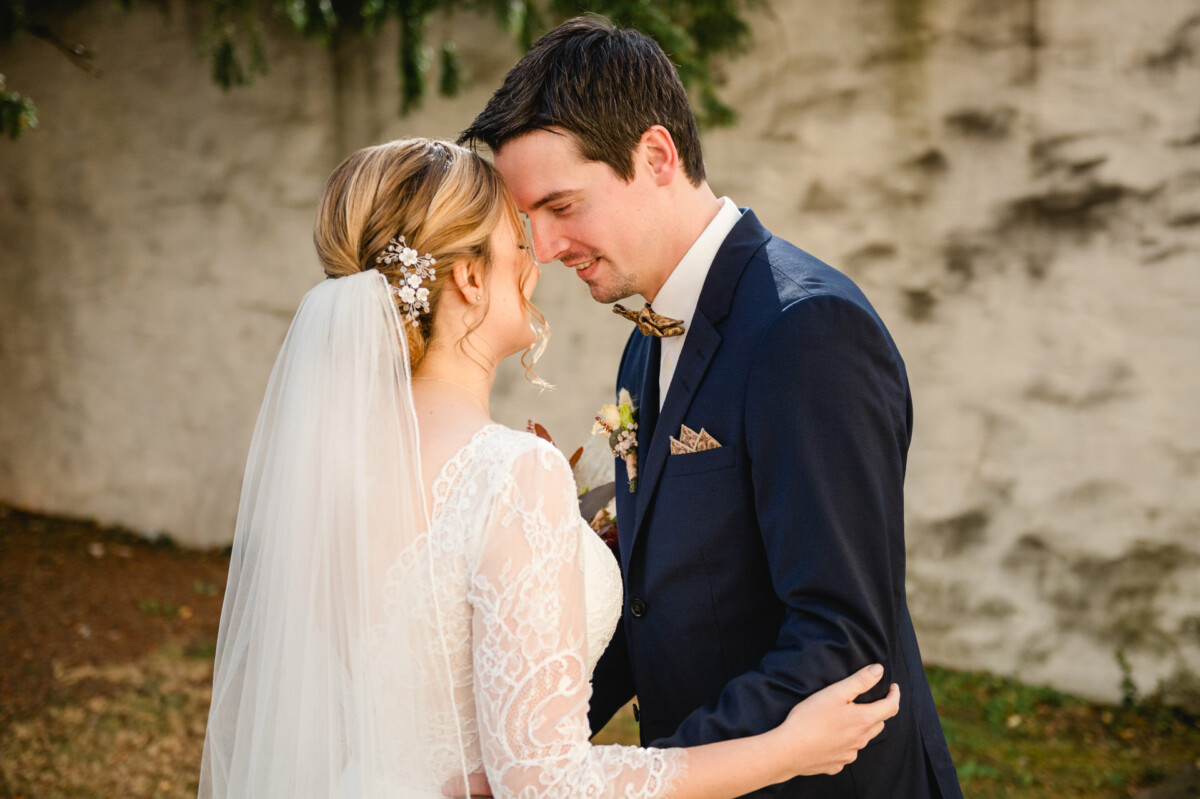 Braut und Bräutigam umarmen sich im Freien. Sie trägt ein weißes Spitzenkleid mit Schleier, er einen marineblauen Anzug mit Fliege.