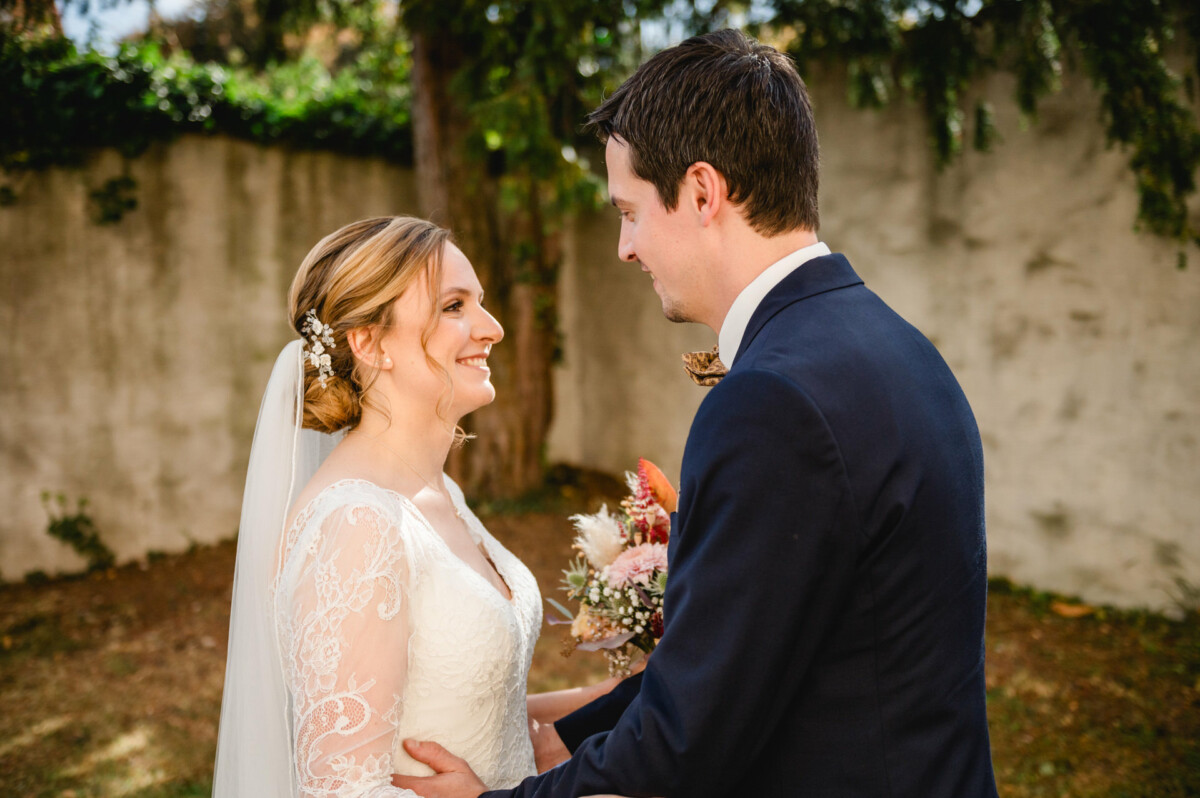 Braut und Bräutigam stehen sich lächelnd im Freien gegenüber. Die Braut hält einen Blumenstrauß in der Hand und trägt ein weißes Spitzenkleid mit Schleier. Im Hintergrund sind eine Mauer und Grünpflanzen zu sehen.