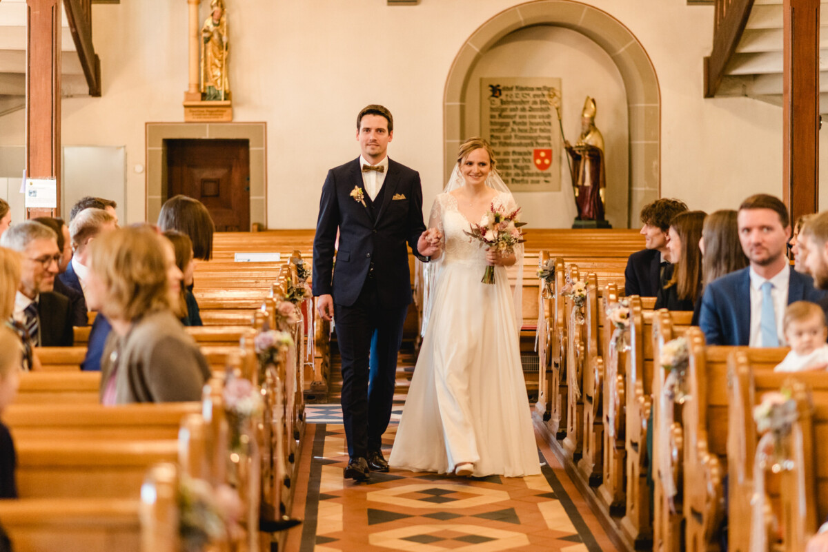 christian-lorenz.net Eine Braut und ein Bräutigam gehen in einer Kirche gemeinsam den Gang entlang, umgeben von sitzenden Gästen.