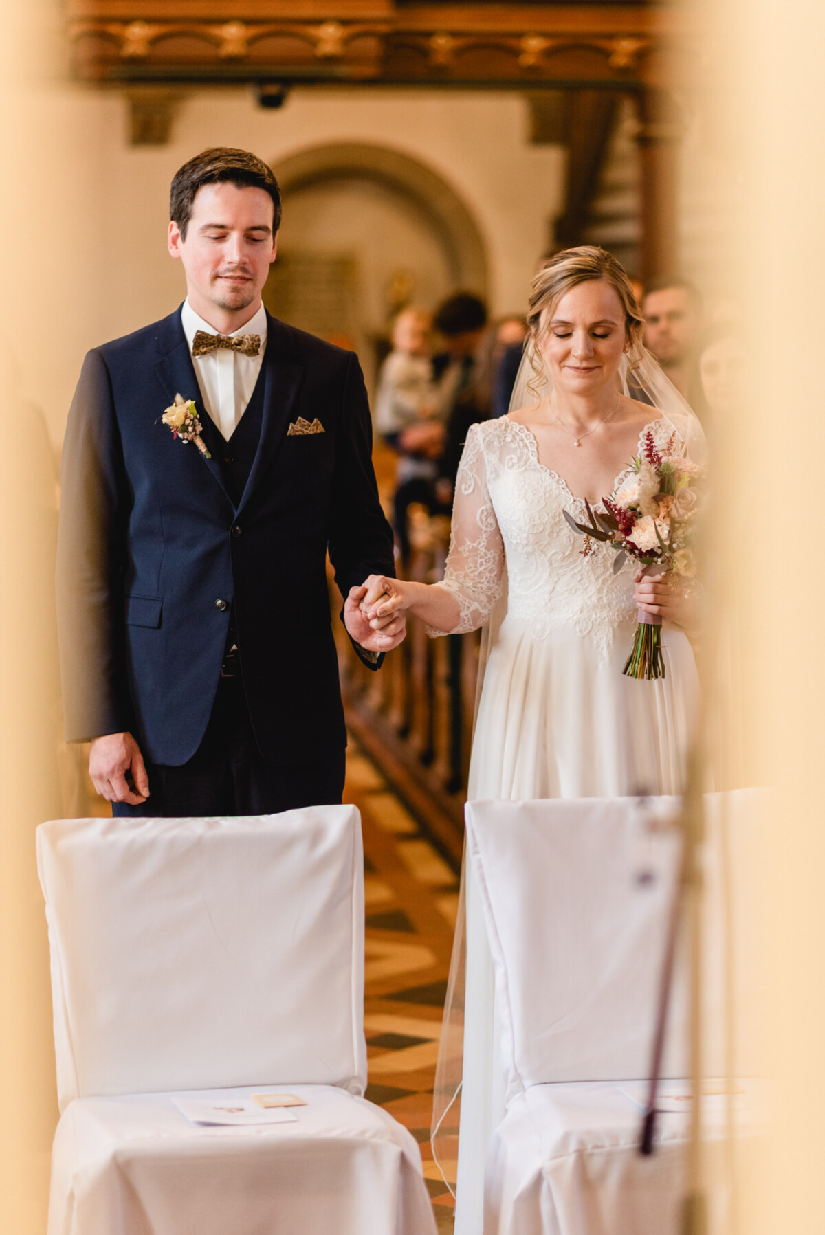 christian-lorenz.net Ein Paar hält während einer Hochzeitszeremonie Händchen. Der Mann trägt einen Anzug, die Frau ein weißes Kleid und hält einen Blumenstrauß in der Hand. Sie stehen in einem Raum mit Stühlen und Holzboden.