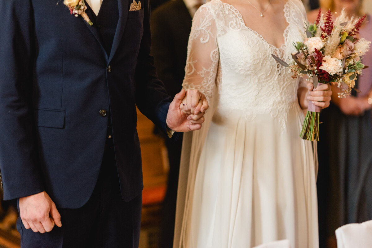 christian-lorenz.net Ein Paar hält während einer Hochzeitszeremonie Händchen. Die Braut trägt ein Spitzenkleid und hält einen Blumenstrauß, während der Bräutigam einen marineblauen Anzug trägt.