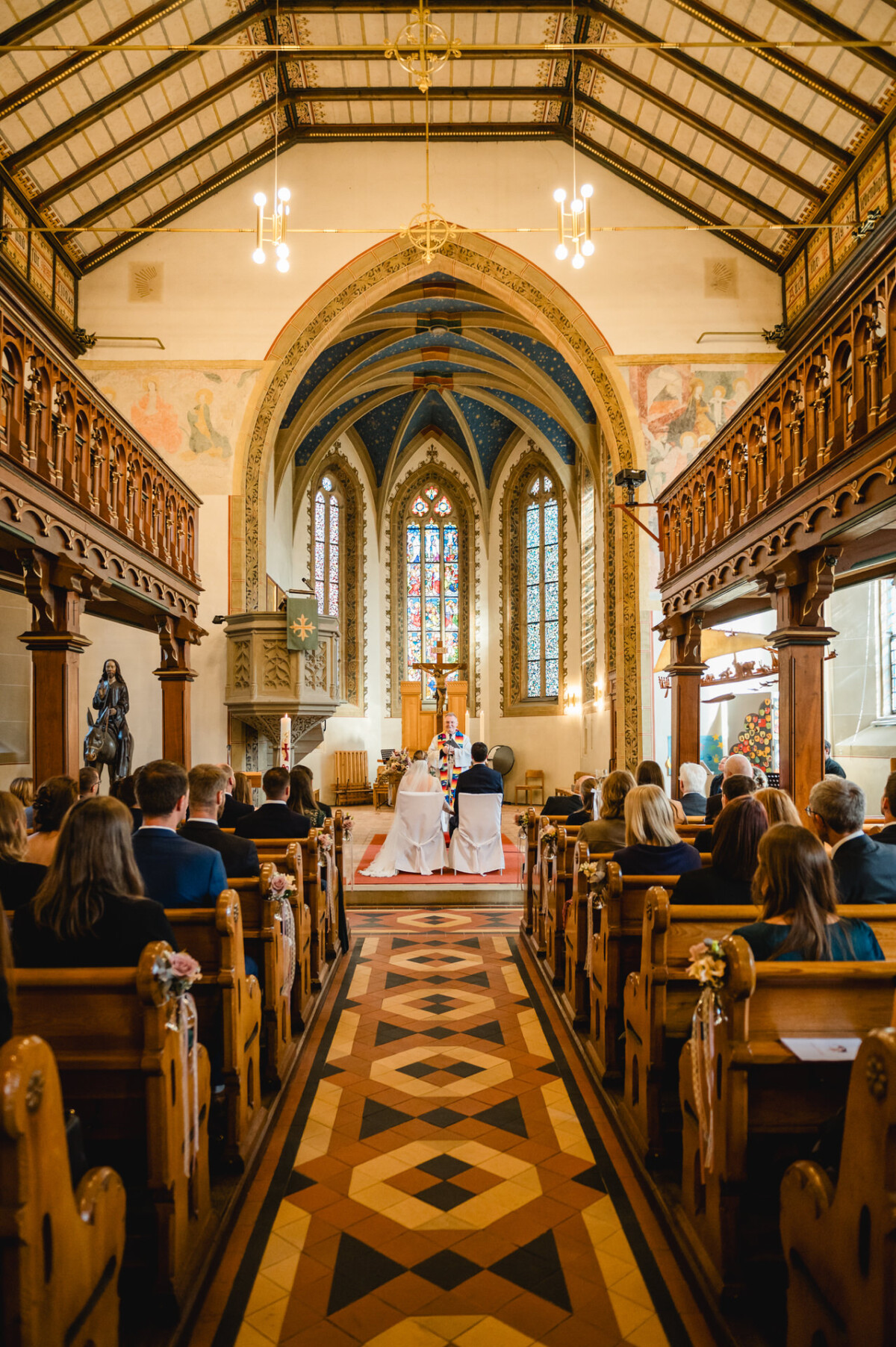 Innenansicht einer Kirche während einer Hochzeitszeremonie. Das Paar sitzt am Altar. Die Gäste sitzen auf Holzbänken, und die Decke und die Buntglasfenster sind sichtbar.
