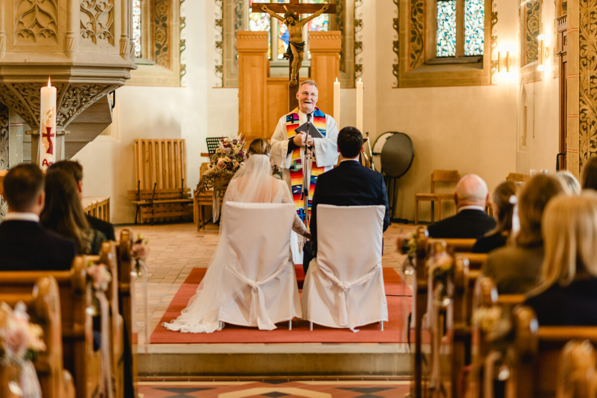 christian-lorenz.net Ein Priester führt in einer Kirche eine Hochzeitszeremonie durch. Braut und Bräutigam sitzen mit Blick auf den Altar, während die Gäste in Holzbänken Platz nehmen.