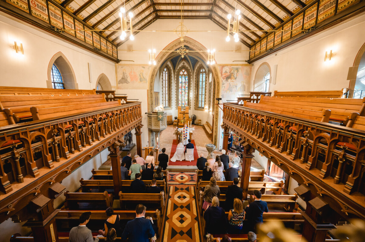 Innenansicht einer Kirche während einer Hochzeitszeremonie mit Holzbänken, dekorativer Decke und Teilnehmern, die dem Altar zugewandt sind.