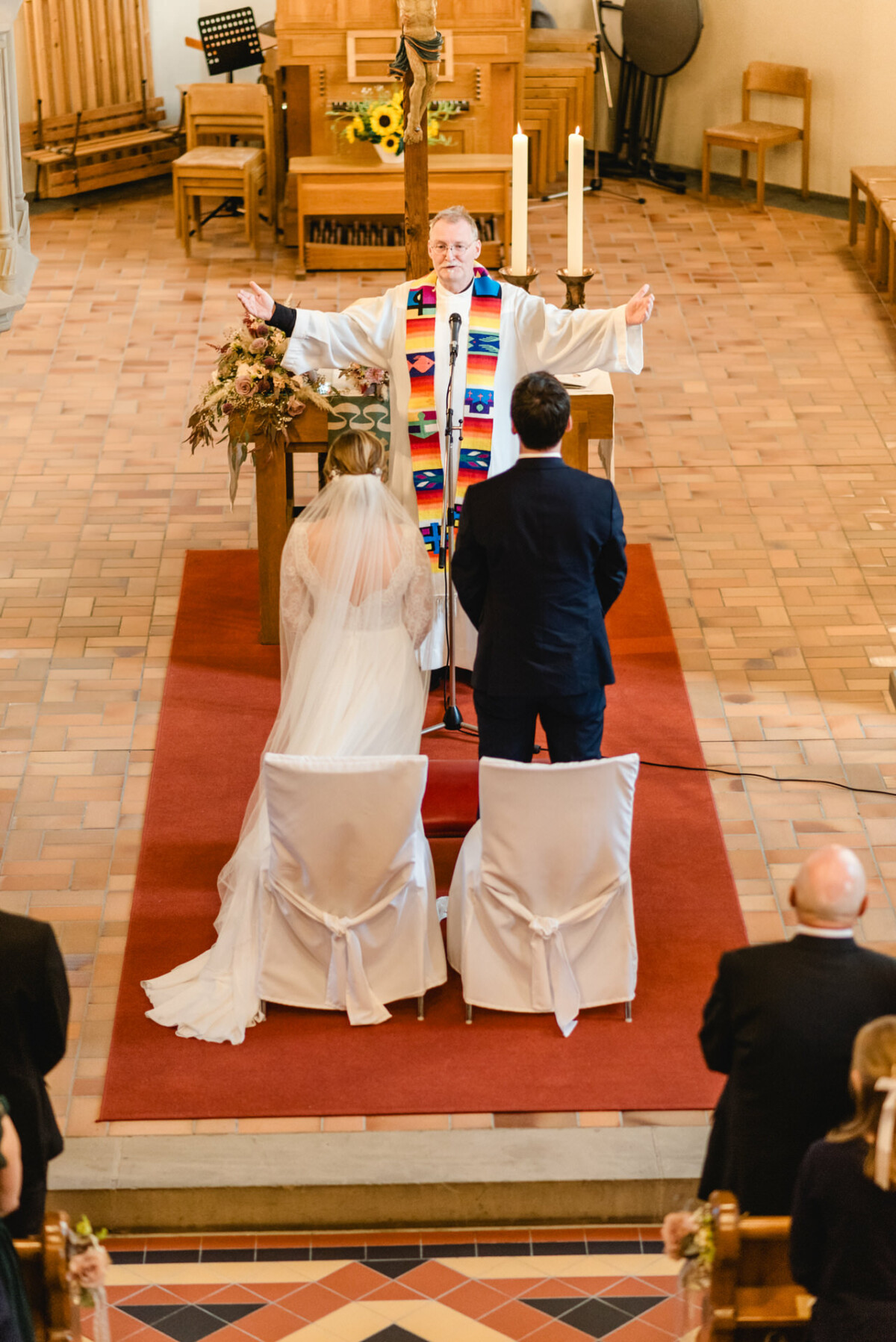 christian-lorenz.net Ein Paar heiratet in einer Kirche. Eine Person mit einer bunten Stola führt die Zeremonie am Altar durch. Die Gäste sitzen in den Kirchenbänken.