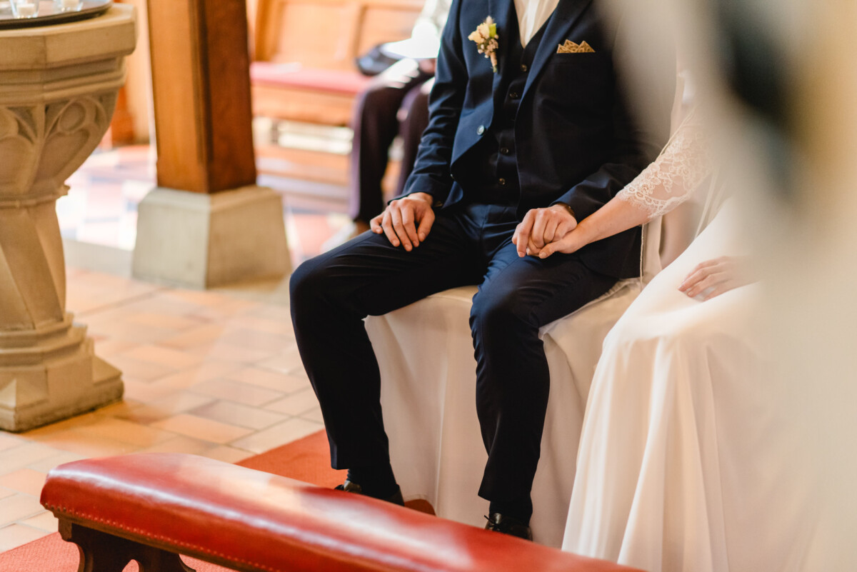 christian-lorenz.net Ein Paar sitzt in einer Kirche und hält sich an den Händen. Der Mann trägt einen dunklen Anzug, die Frau ein weißes Kleid. Sie sitzen auf einer Bank mit Holzelementen im Hintergrund.