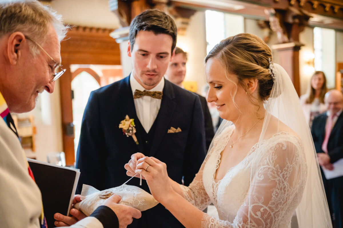 Braut und Bräutigam bei einer Hochzeitszeremonie. Die Braut legt einen Ring auf ein Kissen, das ein Geistlicher hält. Sie befinden sich in einem dekorierten Saal, während die Gäste zuschauen.