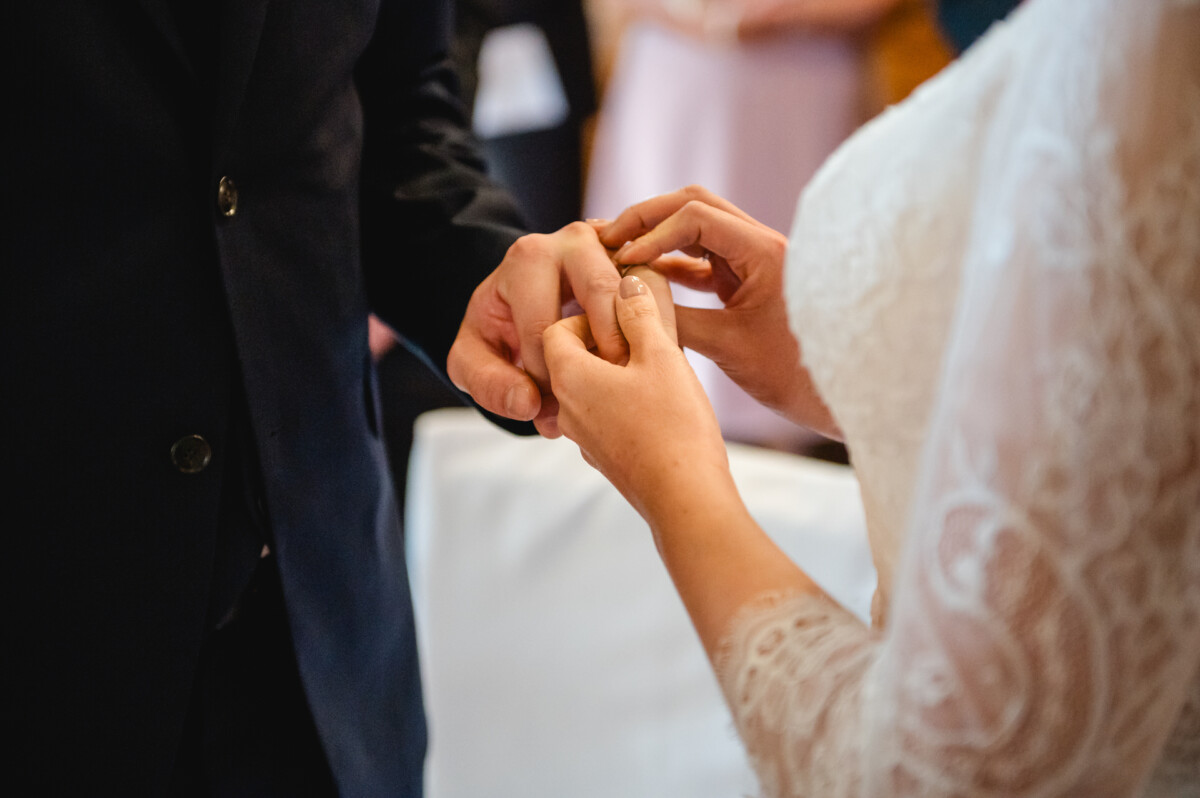 Während einer Hochzeitszeremonie steckt eine Person einer anderen Person einen Ring an den Finger. Beide tragen formelle Kleidung.