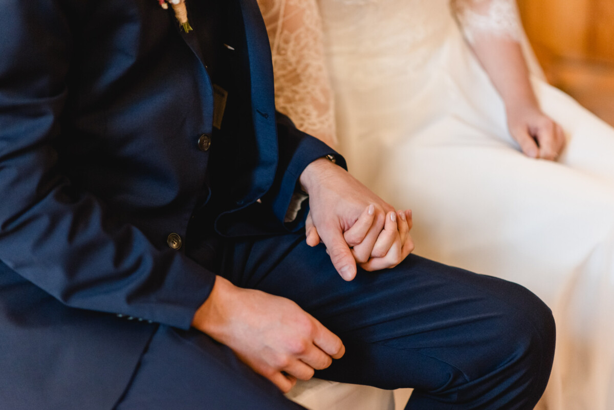christian-lorenz.net Ein Paar sitzt eng beieinander und hält Händchen. Die Person links trägt einen dunklen Anzug, die Person rechts ein helles Kleid.