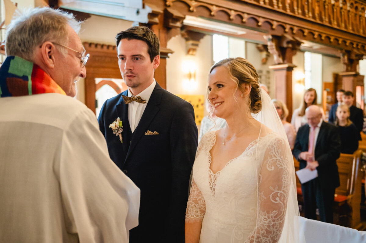 Ein Paar steht in einer Kirche vor dem Altar und blickt einem Geistlichen zu. Die Braut trägt ein weißes Kleid, der Bräutigam einen dunklen Anzug.
