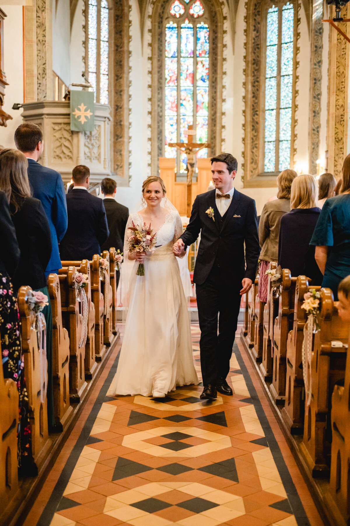 Ein Brautpaar geht den Gang einer mit Gästen gefüllten Kirche entlang, im Hintergrund sind Buntglasfenster zu sehen.