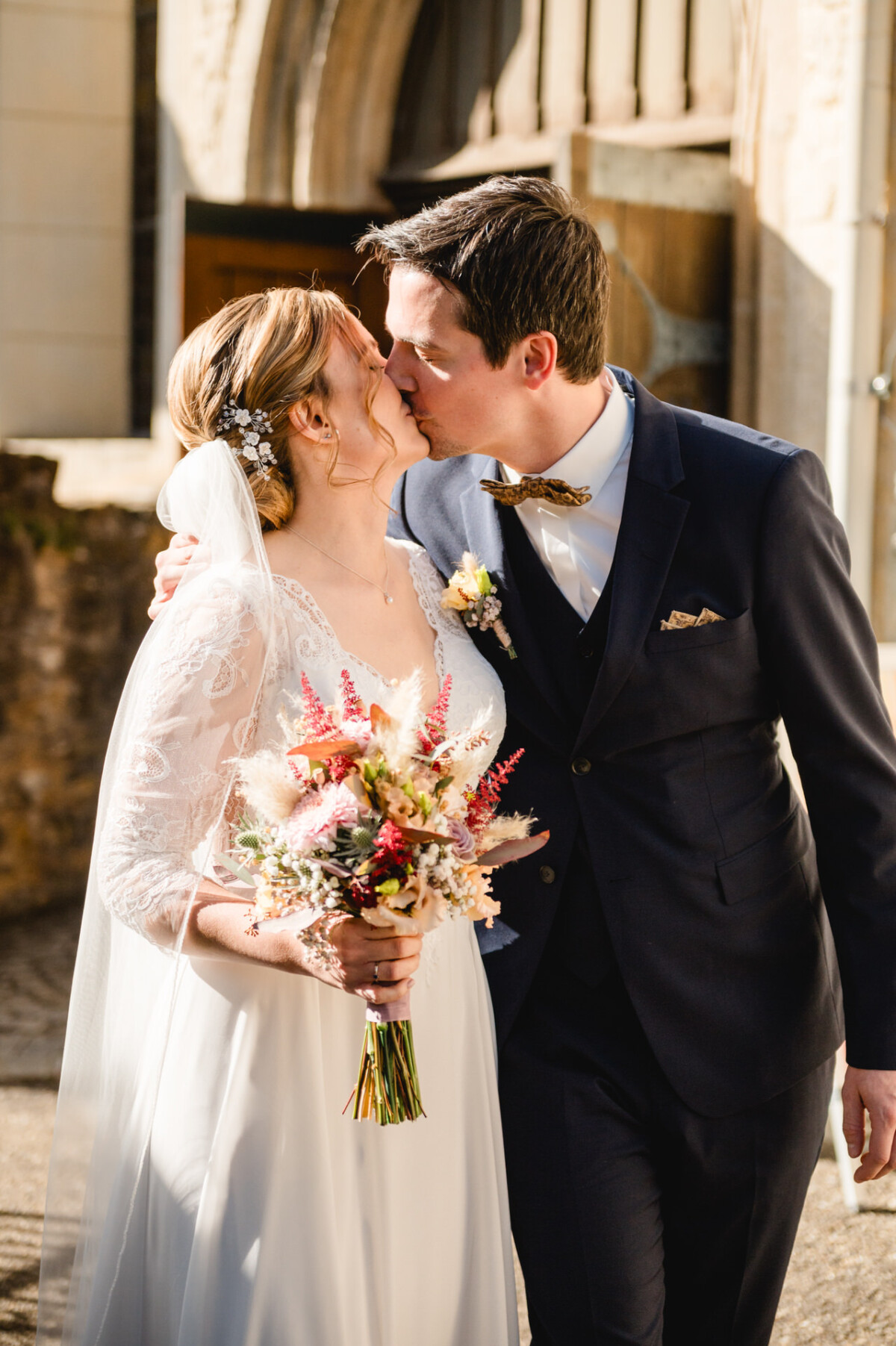 Draußen küssen sich Braut und Bräutigam. Die Braut hält einen Blumenstrauß in der Hand. Beide tragen formelle Hochzeitskleidung.