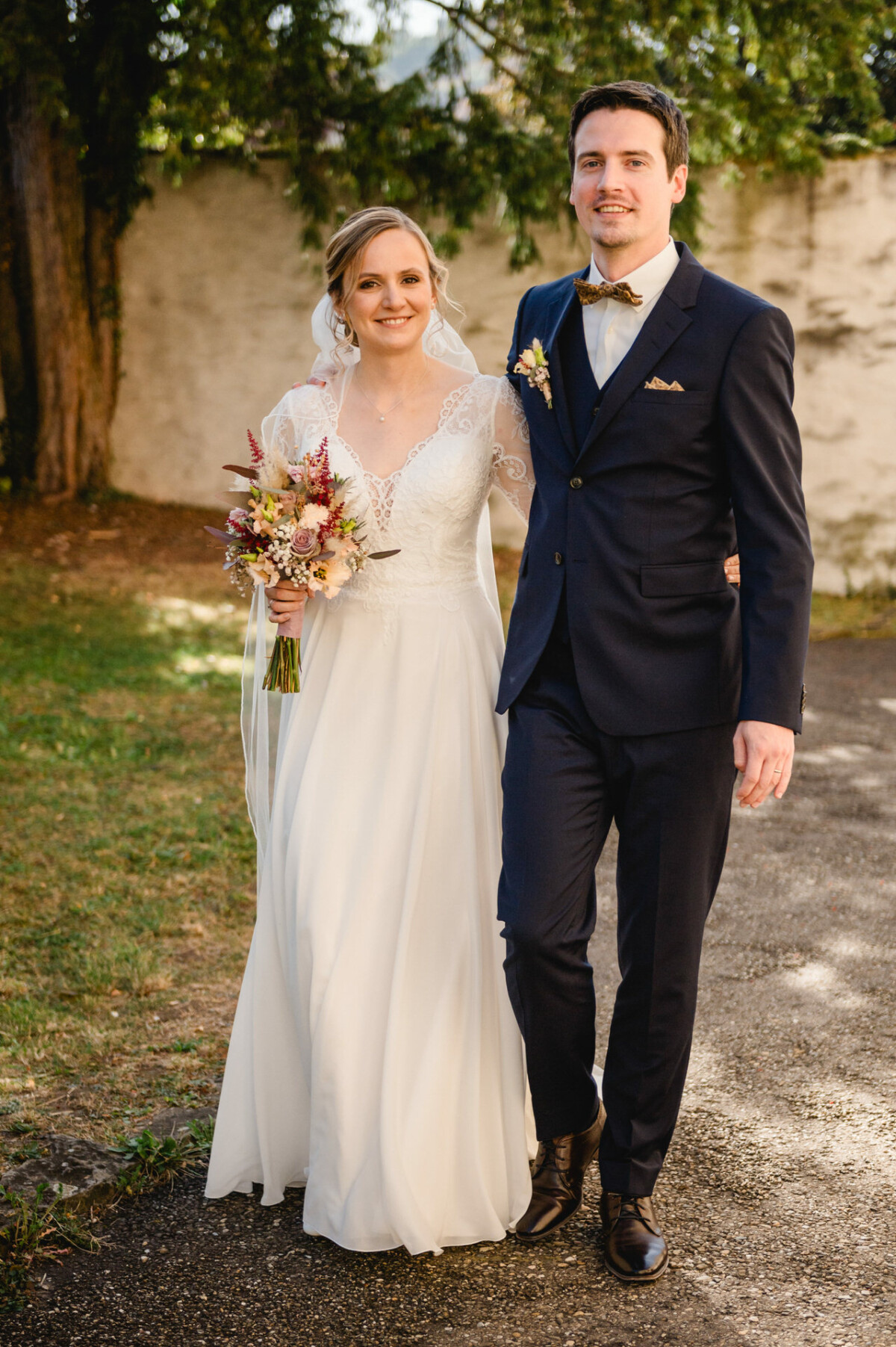 Braut und Bräutigam stehen im Freien auf einem Weg. Die Braut trägt ein weißes Kleid und einen Blumenstrauß in der Hand, der Bräutigam einen schwarzen Anzug mit Fliege. Im Hintergrund sind Bäume und eine Mauer zu sehen.