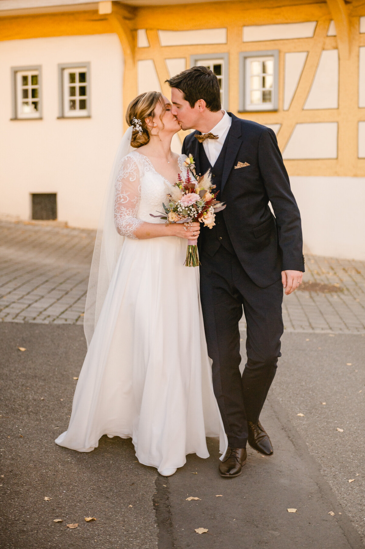 Braut und Bräutigam küssen sich beim Spaziergang im Freien. Die Braut hält einen Blumenstrauß in der Hand und trägt ein Kleid mit Spitzenärmeln, und der Bräutigam trägt einen dunklen Anzug mit Fliege.