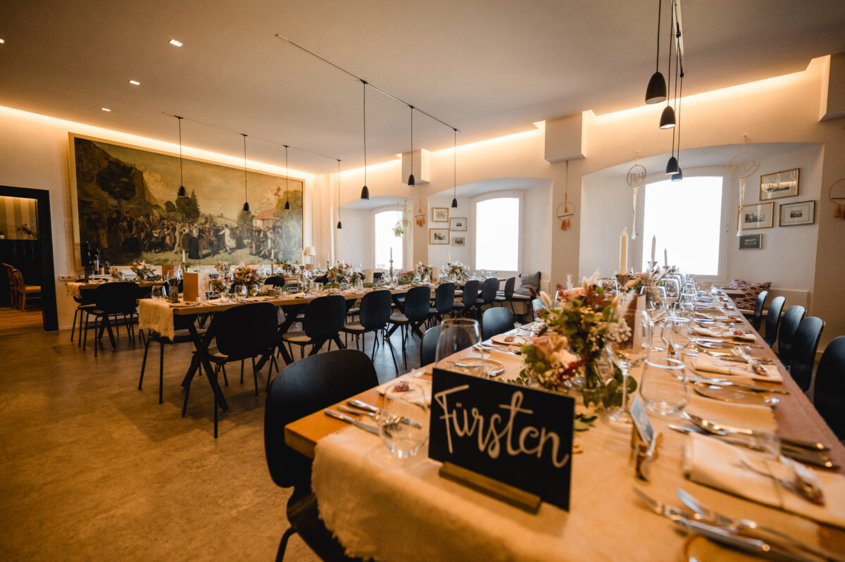 Elegantes Esszimmer für eine formelle Veranstaltung mit langen Tischen, dekorativen Blumenarrangements, Kerzen und schwarzen Stühlen. An der Wand hängt ein großes Wandgemälde.