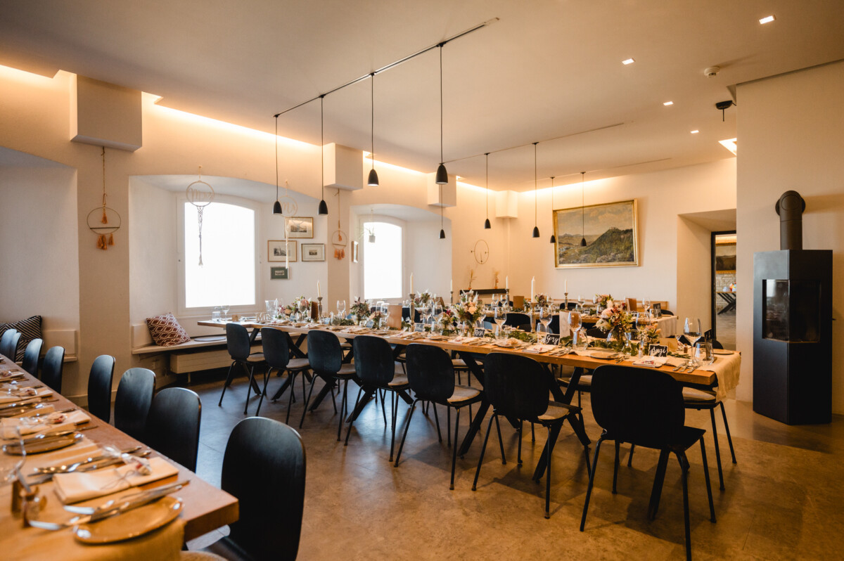 Ein langer Esstisch, gedeckt für eine Veranstaltung in einem hellen, modern eingerichteten Raum. Der Tisch ist mit Blumen geschmückt, Stühle stehen an beiden Seiten. An den Wänden hängen gerahmte Kunstwerke.