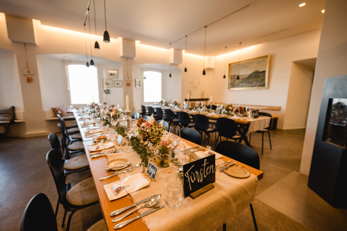 Elegantes Esszimmer in einem geräumigen Raum mit dekorierten langen Tischen, Stühlen und sanfter Beleuchtung. Teller, Gläser und Blumenschmuck schmücken die Tische, und ein „Reserviert“-Schild ist sichtbar.