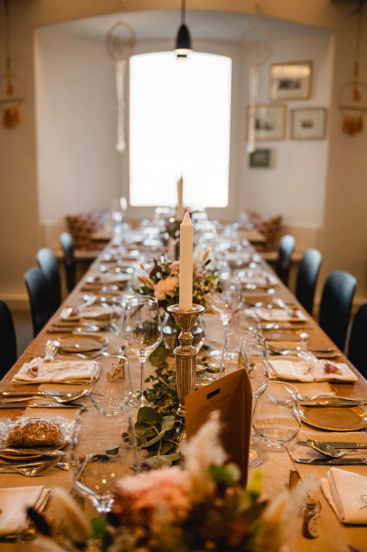 Langer Esstisch mit weißer Tischdecke, gedeckt mit Tellern, Gläsern, Besteck, einer brennenden Kerze und Blumengestecken in einem sanft beleuchteten Raum.