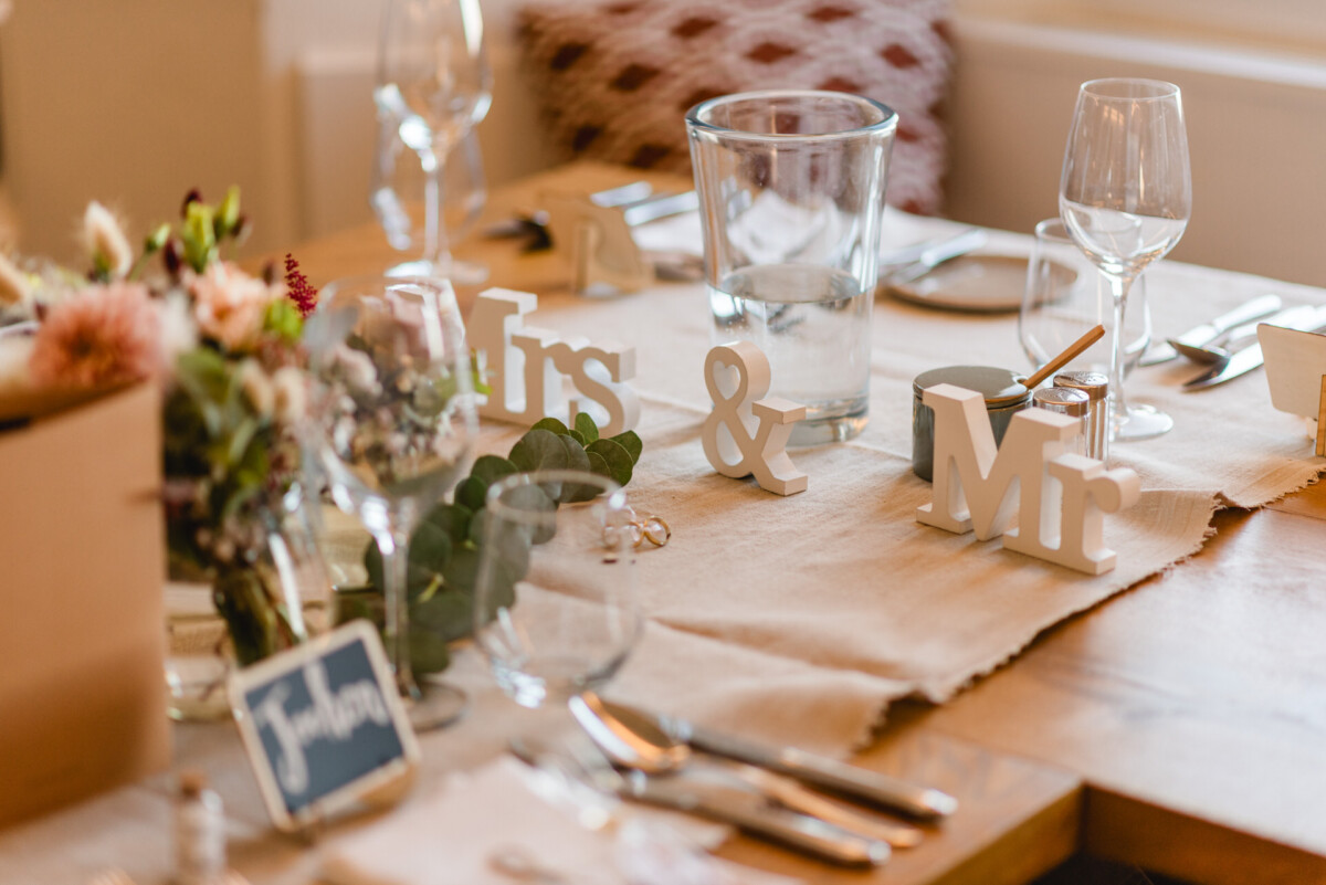 christian-lorenz.net Nahaufnahme einer Hochzeitstafel mit „Mrs & Mr“-Schild, Gläsern, Besteck und Blumendekorationen auf einem Holztisch.