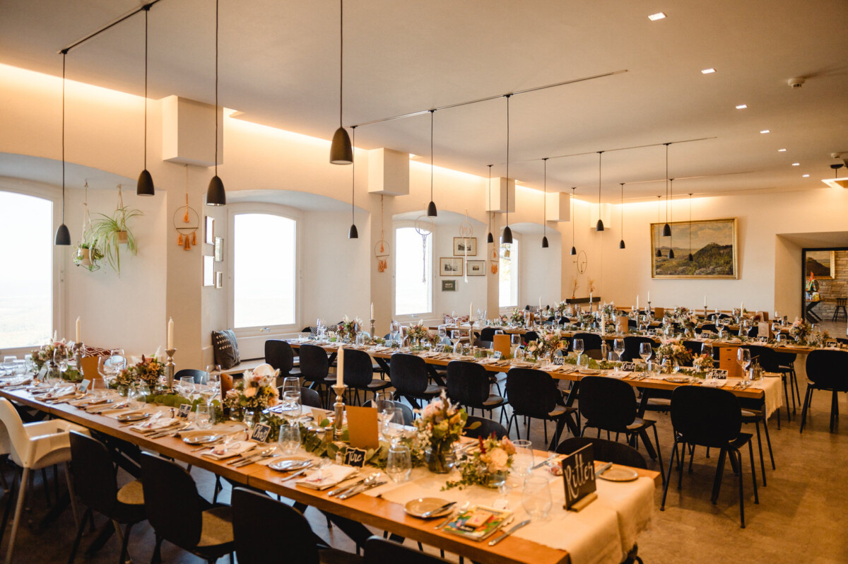 Ein großer Essbereich ist für eine Veranstaltung mit langen Tischen, geschmückt mit Blumengestecken, Kerzen und Geschirr, eingerichtet. Schwarze Stühle umgeben die Tische, und von der Decke hängen Pendelleuchten.