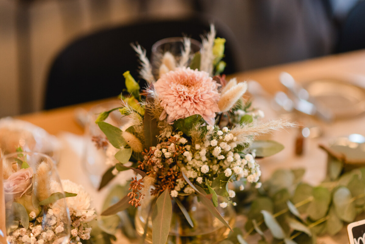 christian-lorenz.net Ein florales Tafelaufsatz mit rosa Blumen, Schleierkraut und Grünpflanzen steht auf einem Esstisch, der mit Besteck und Tellern gedeckt ist.