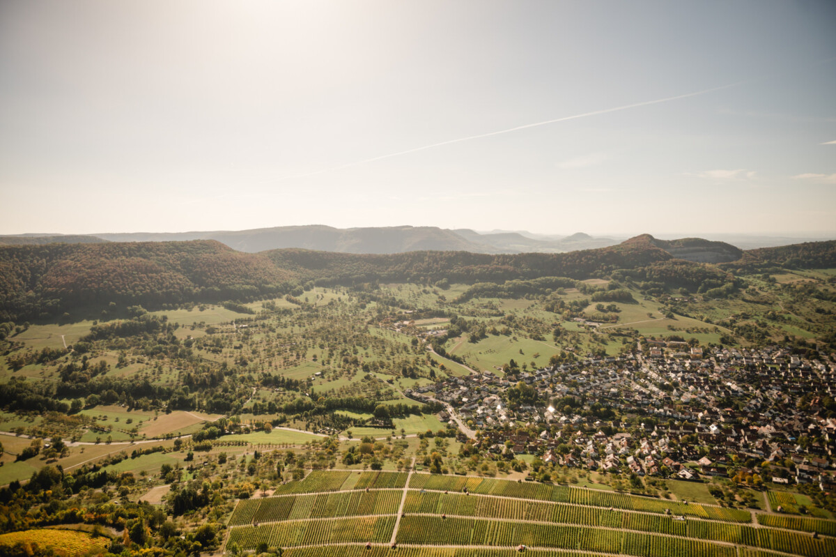 Luftaufnahme einer ländlichen Landschaft mit Feldern, einer kleinen Stadt und umliegenden Hügeln unter klarem Himmel.
