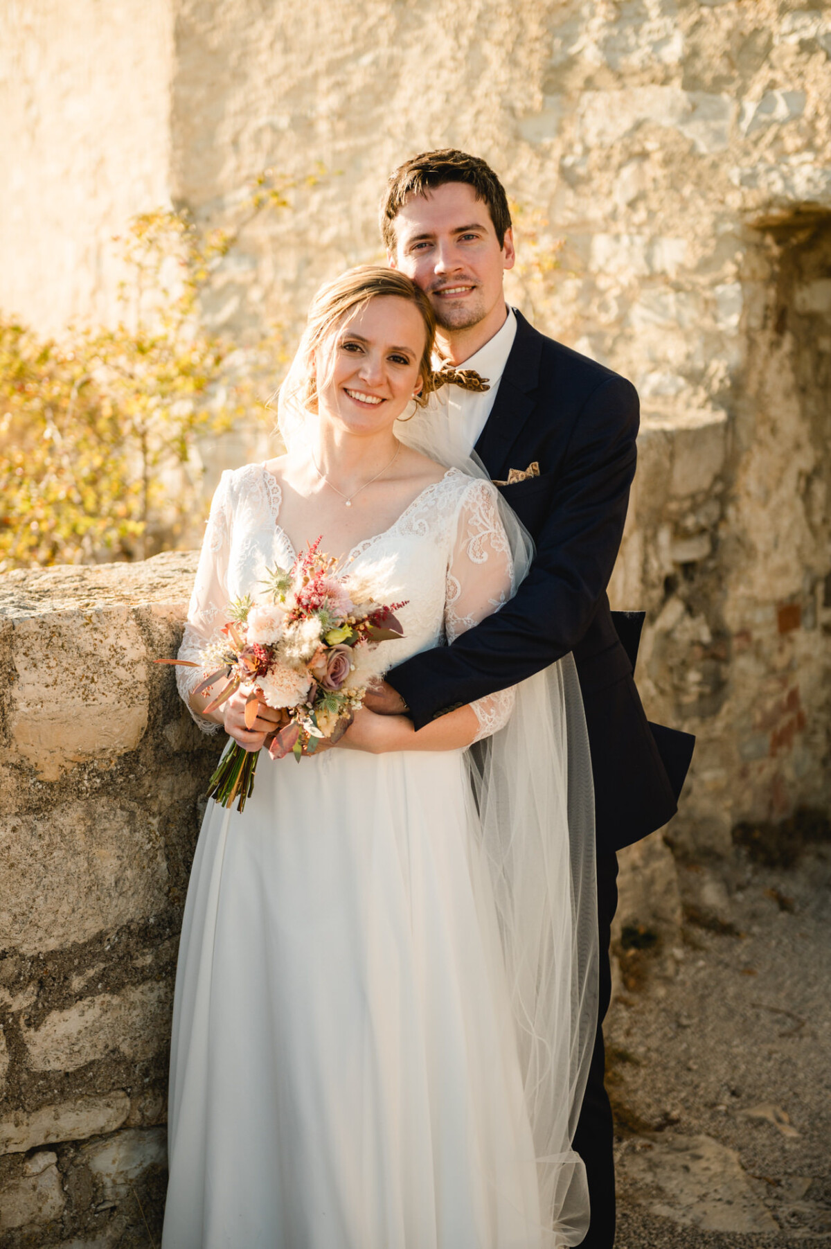 Braut und Bräutigam posieren gemeinsam im Freien. Die Braut hält einen Blumenstrauß in der Hand und trägt ein weißes Kleid mit Spitzenärmeln. Der Bräutigam steht in dunklem Anzug und Krawatte hinter ihr. Sie stehen vor einer Steinmauer.