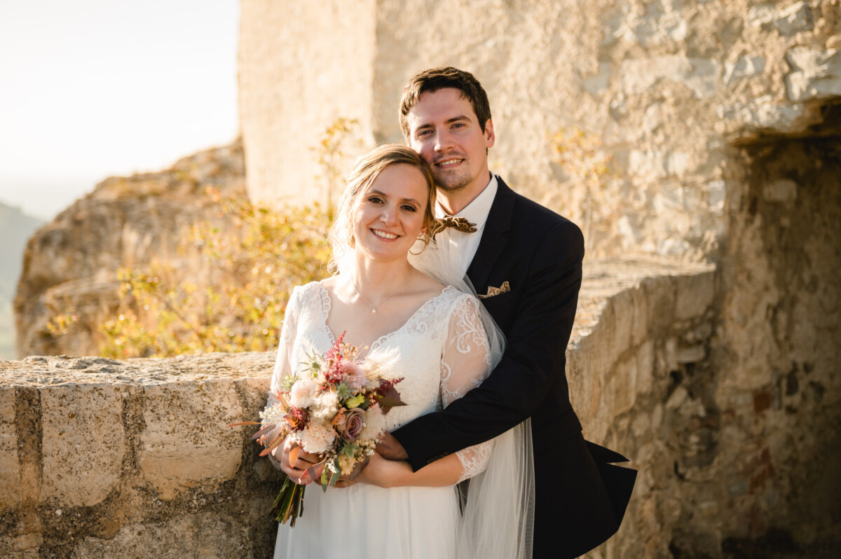 Braut und Bräutigam posieren im Hochzeitskleid im Freien vor einer Steinmauer. Die Braut hält einen Blumenstrauß und beide lächeln in die Kamera.