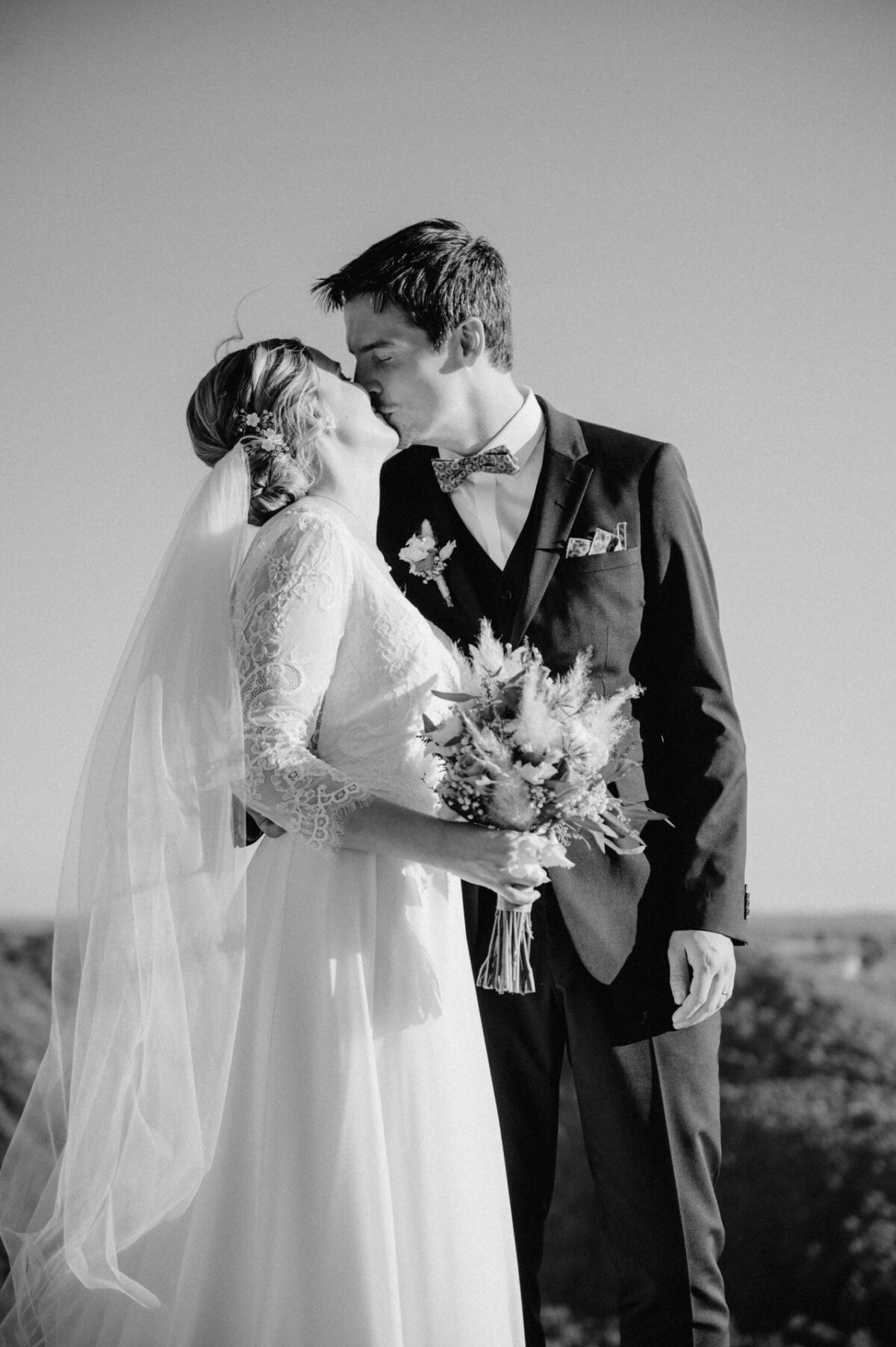 Braut und Bräutigam küssen sich im Freien, sie hält einen Blumenstrauß, beide in Abendgarderobe. Schwarzweißfoto.