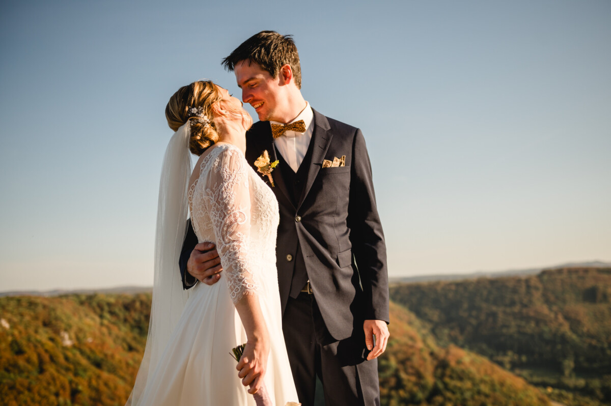 Braut und Bräutigam stehen dicht beieinander und lächeln vor der Kulisse sanfter Hügel unter einem klaren blauen Himmel.