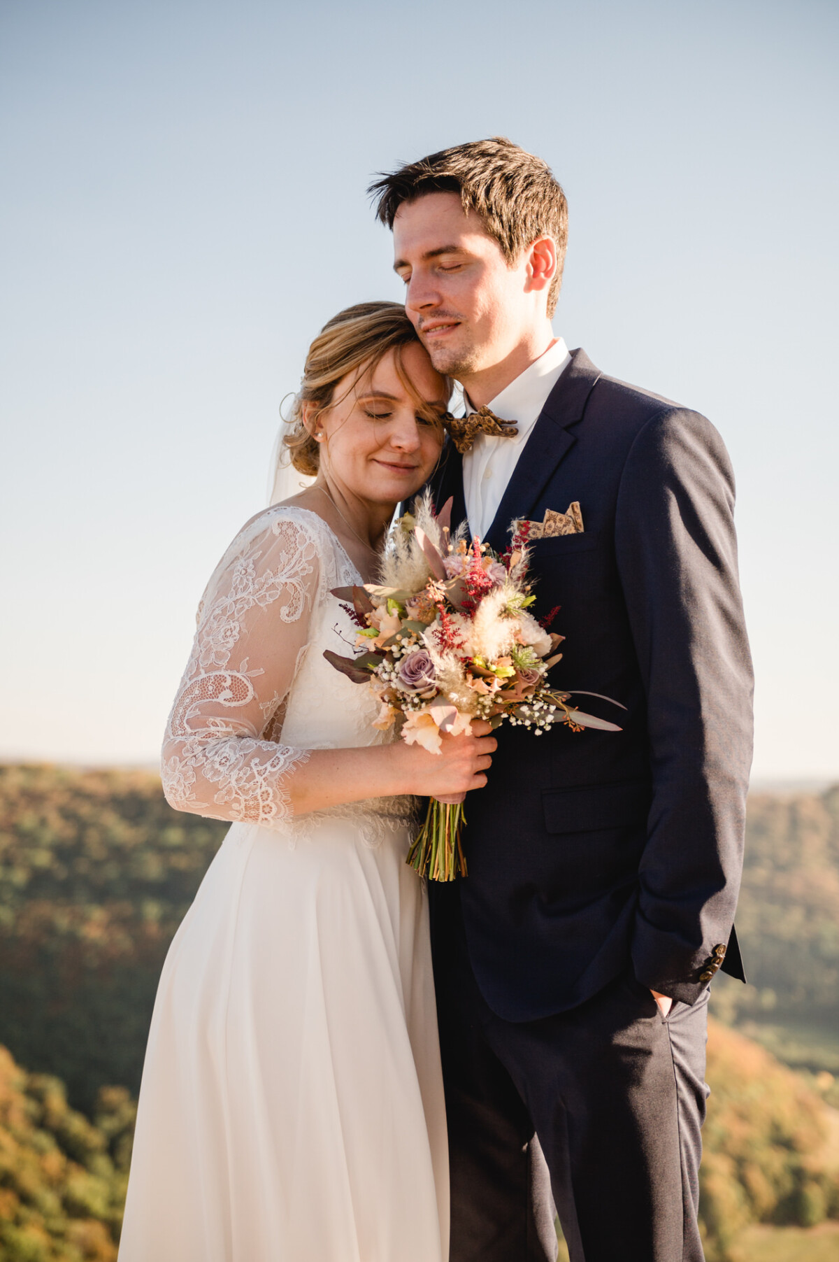 Braut und Bräutigam umarmen sich im Freien, die Braut hält einen Blumenstrauß. Sie stehen vor einer malerischen, sonnenbeschienenen Kulisse.