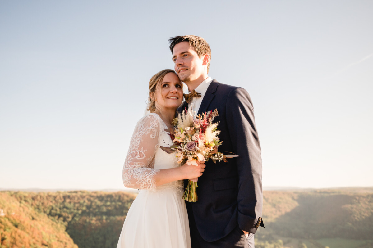 Braut und Bräutigam stehen im Freien mit einer malerischen Landschaft im Hintergrund. Die Braut hält einen Blumenstrauß in der Hand und trägt ein weißes Kleid, während der Bräutigam einen Anzug trägt.