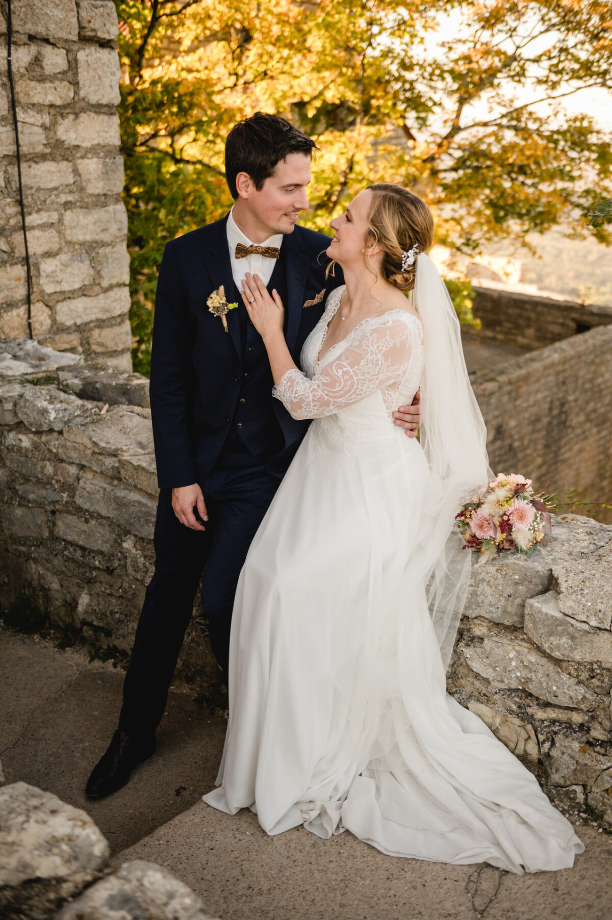 Eine Braut und ein Bräutigam im Hochzeitskleid lächeln einander an, während sie auf einer Steinmauer sitzen, umgeben von Herbstlaub.