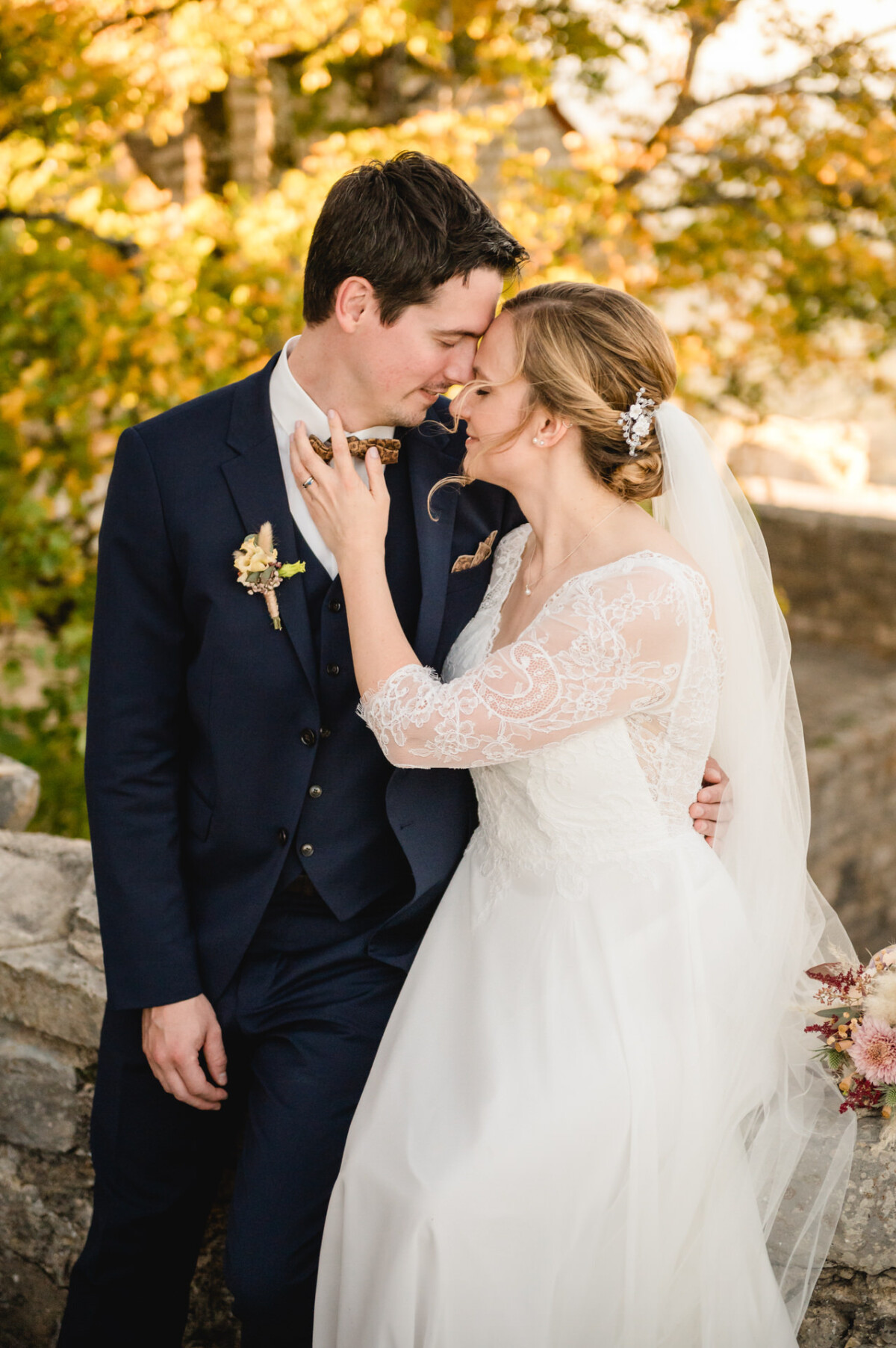 Braut und Bräutigam teilen einen zärtlichen Moment im Freien. Die Braut trägt ein weißes Kleid und einen Schleier, der Bräutigam einen marineblauen Anzug. Umgeben von herbstlichem Laub berühren sie sanft ihre Stirn.