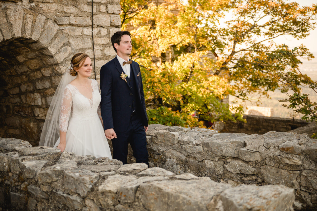 Eine Braut und ein Bräutigam halten Händchen und stehen auf einem Steinpfad, umgeben von Herbstbäumen und Steinarchitektur.