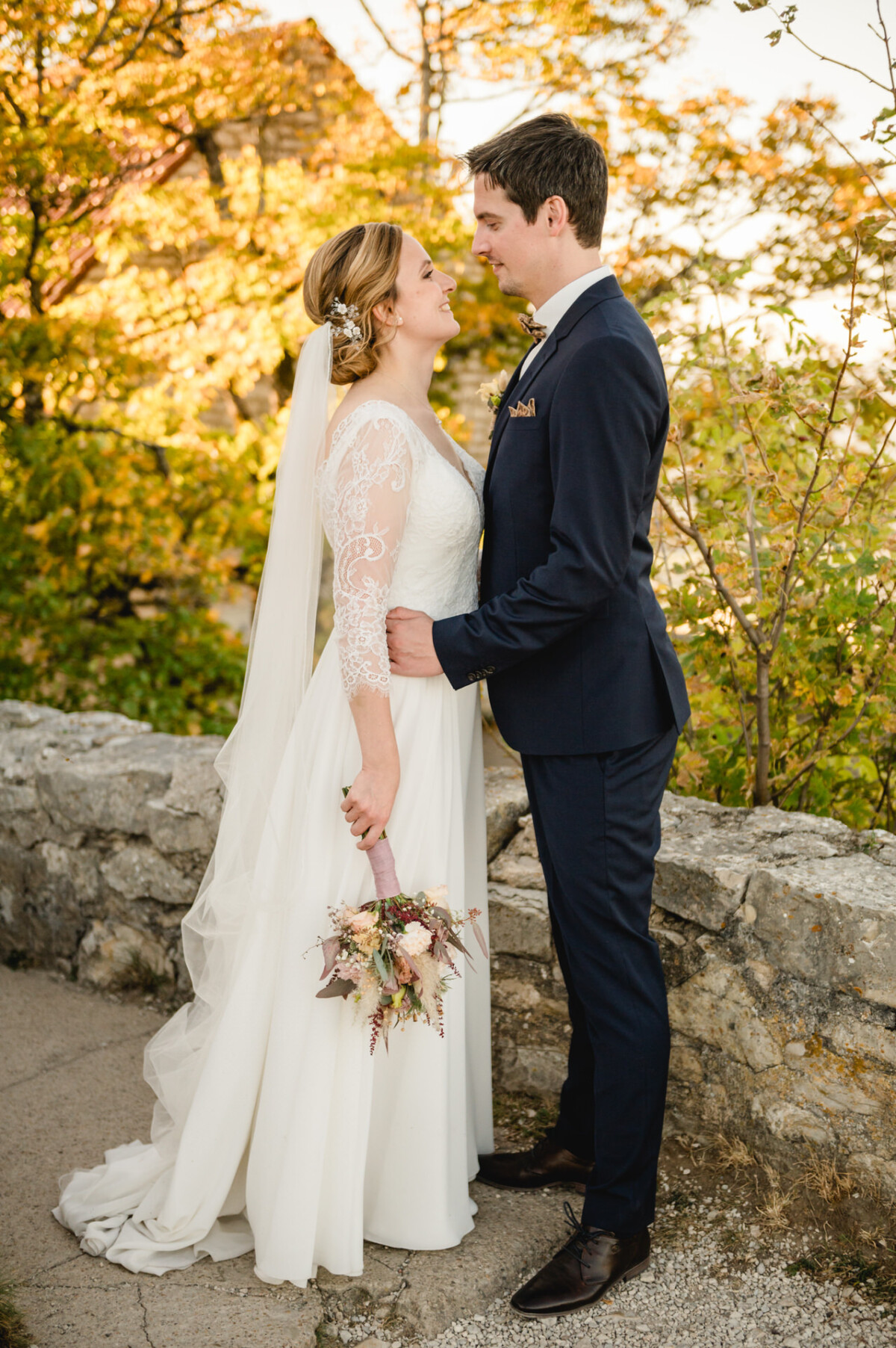 Braut im weißen Kleid und Bräutigam im marineblauen Anzug stehen sich lächelnd gegenüber. Sie hält einen Blumenstrauß in der Hand. Im Hintergrund sind Grünflächen und eine Steinmauer zu sehen.