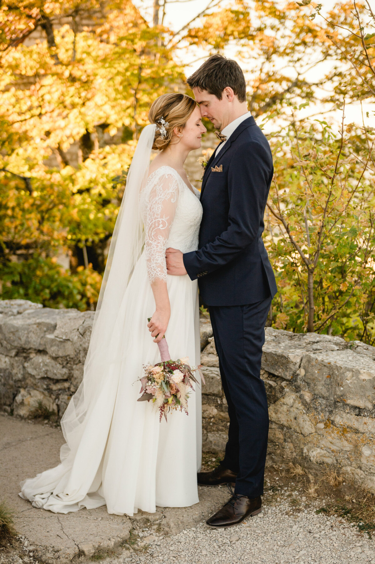 Braut und Bräutigam stehen einander gegenüber, eng aneinandergedrängt, im Hintergrund ist herbstliches Laub zu sehen. Die Braut hält einen Blumenstrauß und beide tragen formelle Hochzeitskleidung.